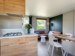 A modern kitchen with wooden cabinets, a gas stove, a dining area with a wooden table and white chairs, and a window with a view of greenery. - Camping 4 étoiles - Parc aquatique - ccbg0cd (Guidel)