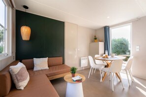 Ein modernes Wohnzimmer mit einem braunen Sofa, einem runden Esstisch mit weißen Stühlen, einer Hängeleuchte und Blick ins Grüne.