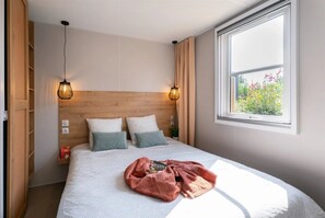 A bedroom with a wooden headboard, two bedside lamps, a window with a view of greenery, and a neatly made bed with pillows and a blanket. - Camping 4 étoiles - Parc aquatique - ccbg0ch (Guidel)