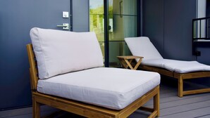 Två träfärgade loungestolar med vita kuddar på en balkong med en glasdörr som leder ut till en trädgård.