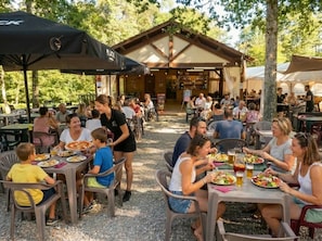 An outdoor dining area with tables and chairs, a covered seating area, and a building in the background. - Camping 3 étoiles - Piscine  - ccbg0ie (Saint-Gérons)