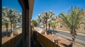 A view from a balcony overlooking a road lined with palm trees, a wooden fence, and a building with a modern design. - Sunset Loft By CanariasGetaway (Mogán)