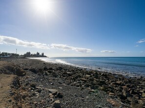 A rocky beach with a clear sky and distant buildings. - Sunset Loft By CanariasGetaway (Mogán)