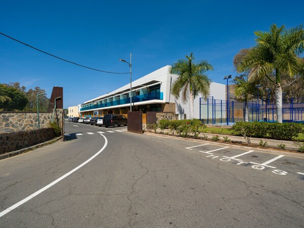 A modern building with a curved driveway, a parking area, and a fenced sports court. - Sunset Loft By CanariasGetaway (Mogán)