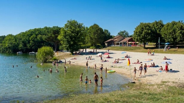 Una playa junto al lago, con gente nadando y tomando el sol, rodeada de árboles y un edificio de techo rojo.