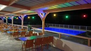 Un área de comedor al aire libre con mesas y sillas de madera, una piscina y luces colgantes.
