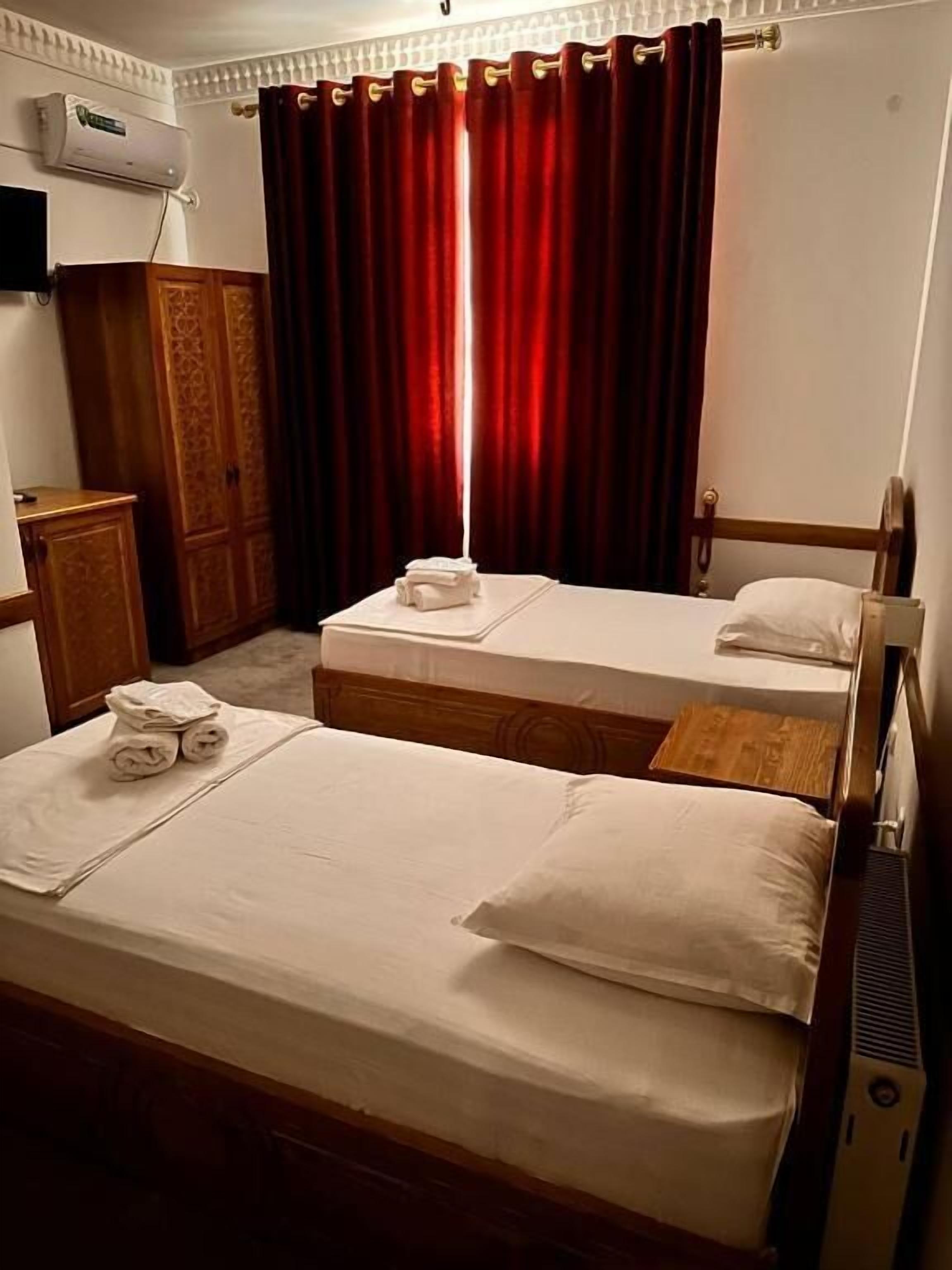 Une chambre d’hôtel avec deux lits simples, une armoire en bois, un téléviseur à écran plat et une fenêtre avec des rideaux rouges.