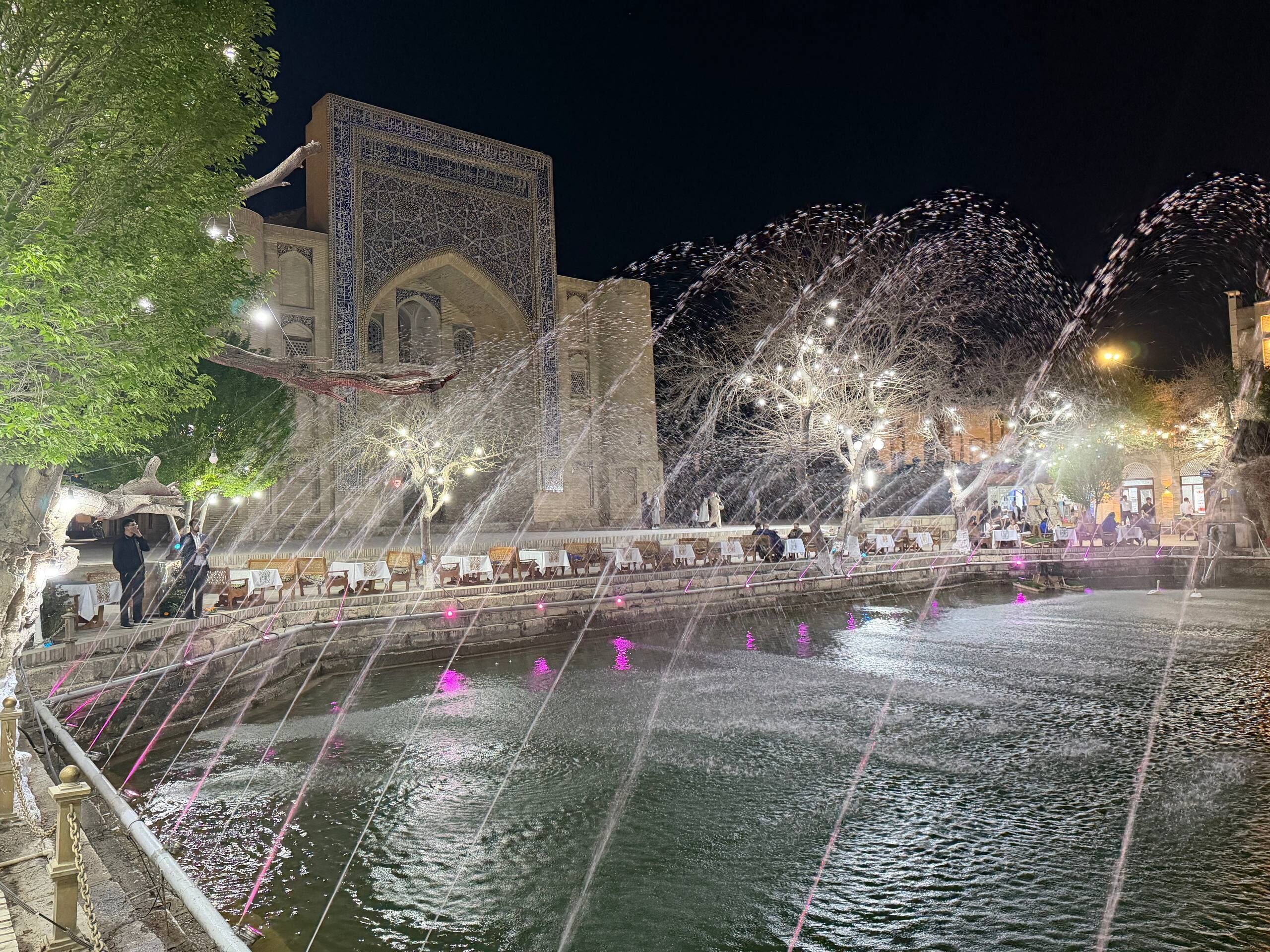 A night scene with a water fountain, a building with intricate architecture, and outdoor seating.