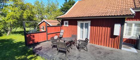 A red wooden building with a tiled roof, a wooden deck with a table and chairs, and a white door.