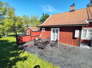 Un bâtiment en bois rouge avec un toit en tuiles, une terrasse en bois avec une table et des chaises, et une porte blanche.