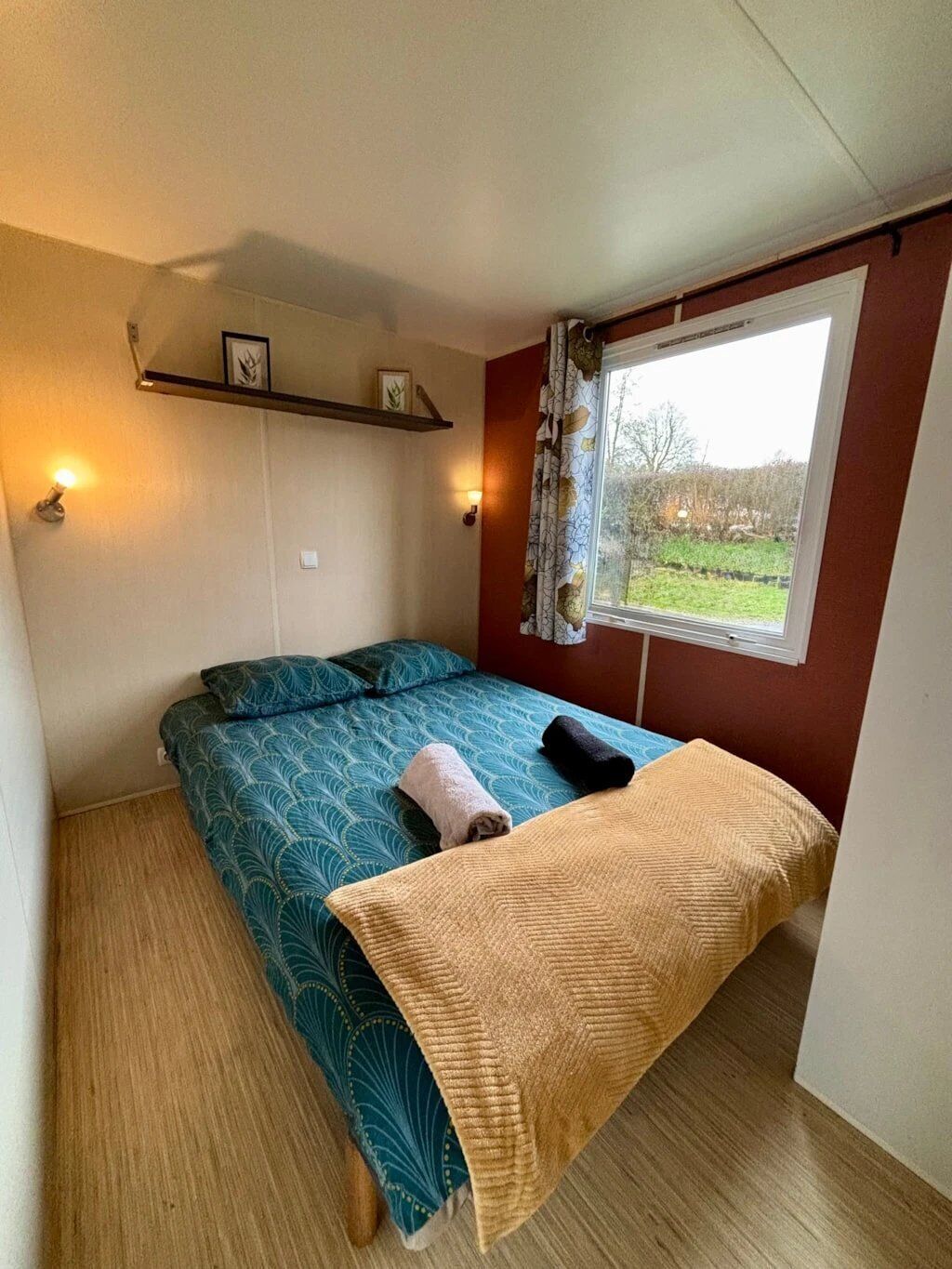 A single bed with patterned bedding, a bedside lamp, a window with curtains, and a shelf with framed pictures.