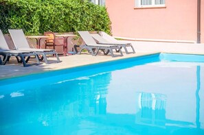 A swimming pool with lounge chairs and a table, surrounded by a pink building and greenery. - Camping 2 étoiles - Piscine  - ccbgbhc (Sospel)