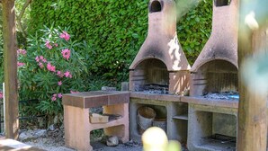 Uma área de cozinha ao ar livre em pedra, com mesa e bancos, rodeada por vegetação e flores cor-de-rosa.