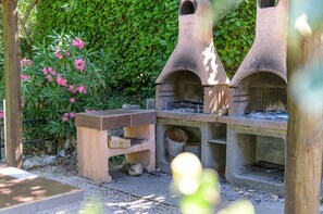 Eine steinerne Außenküche mit Tisch und Bänken, umgeben von Grünpflanzen und rosa Blüten.
