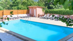 Piscina retangular com espreguiçadeiras ao redor e um pequeno edifício com porta e janelas.