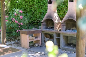 Een buitenkeuken van steen met een tafel en banken, omgeven door groen en roze bloemen.