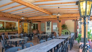 Un café al aire libre con vigas de madera, mesas, sillas y lámparas colgantes.