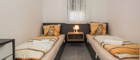 A twin room with two beds, a nightstand with a lamp, a window with sheer curtains, and wall-mounted pictures.
