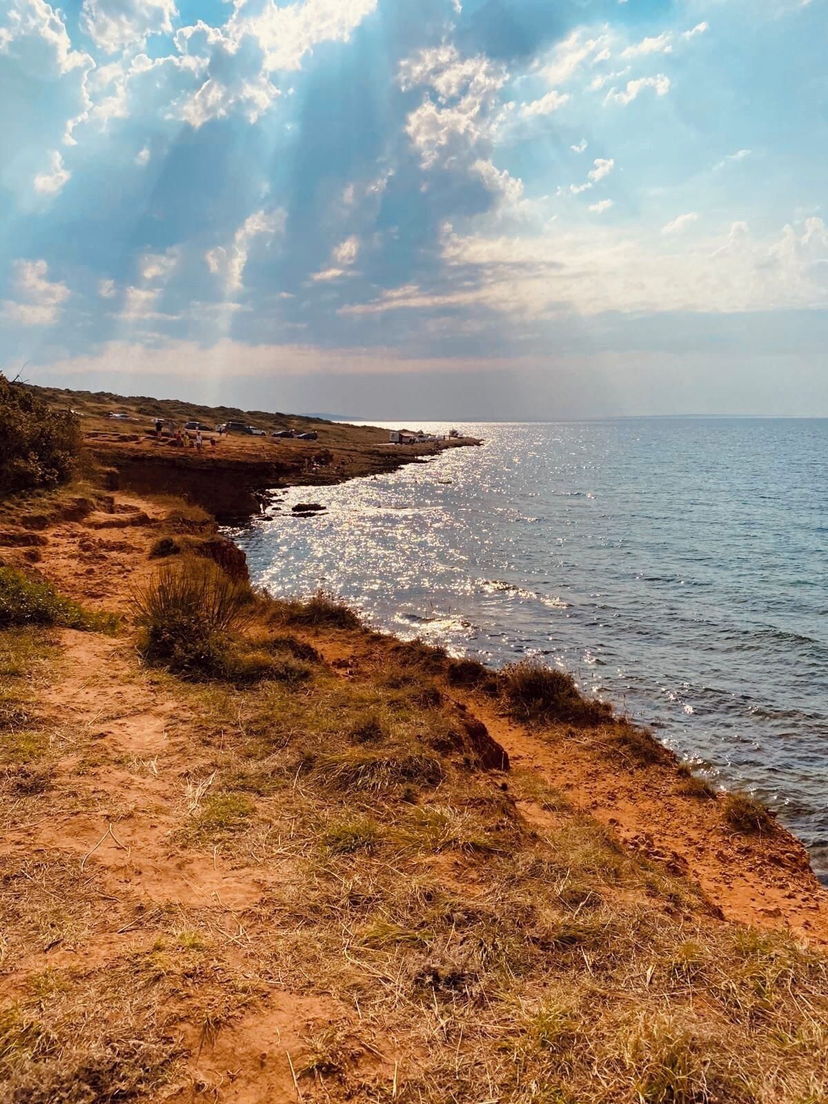 A coastal landscape with a dirt path leading to the sea, under a partly cloudy sky.