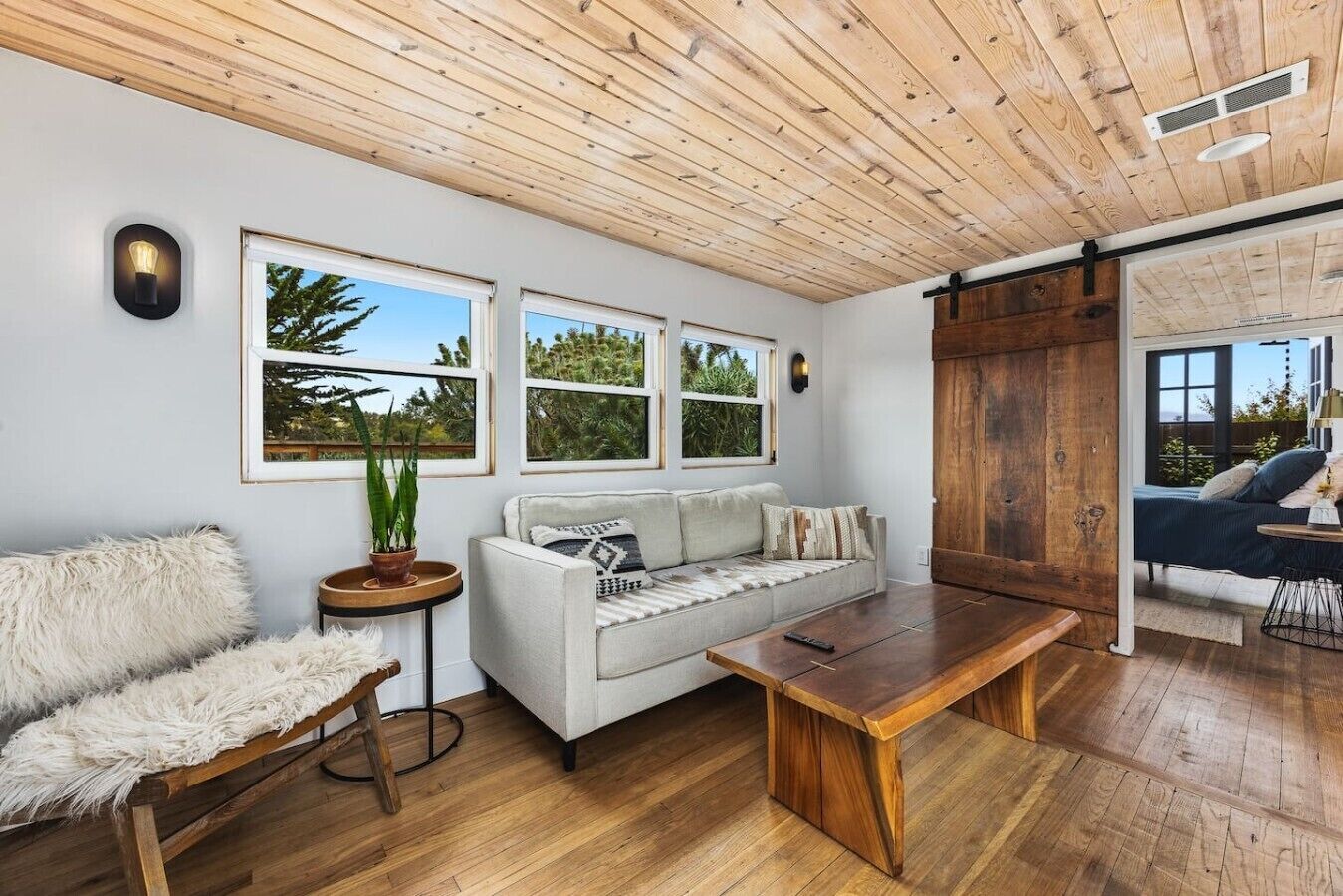 A room with a wooden ceiling, a white sofa, a wooden coffee table, and a large window with a view of trees.