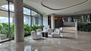 A spacious lobby with a modern design, featuring a seating area with white chairs, a marble floor, and large windows offering a view of greenery outside. - MODERN APT WITH SEA VIEW AND SWIMMING POOLS 1 BEDROOM WITH BALCONY. MANZANILLO BEACH. (CARTAGENA)
