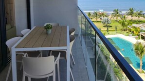 A balcony with a wooden table and chairs, a glass railing, and a view of the ocean and a pool. - MODERN APT WITH SEA VIEW AND SWIMMING POOLS 1 BEDROOM WITH BALCONY. MANZANILLO BEACH. (CARTAGENA)
