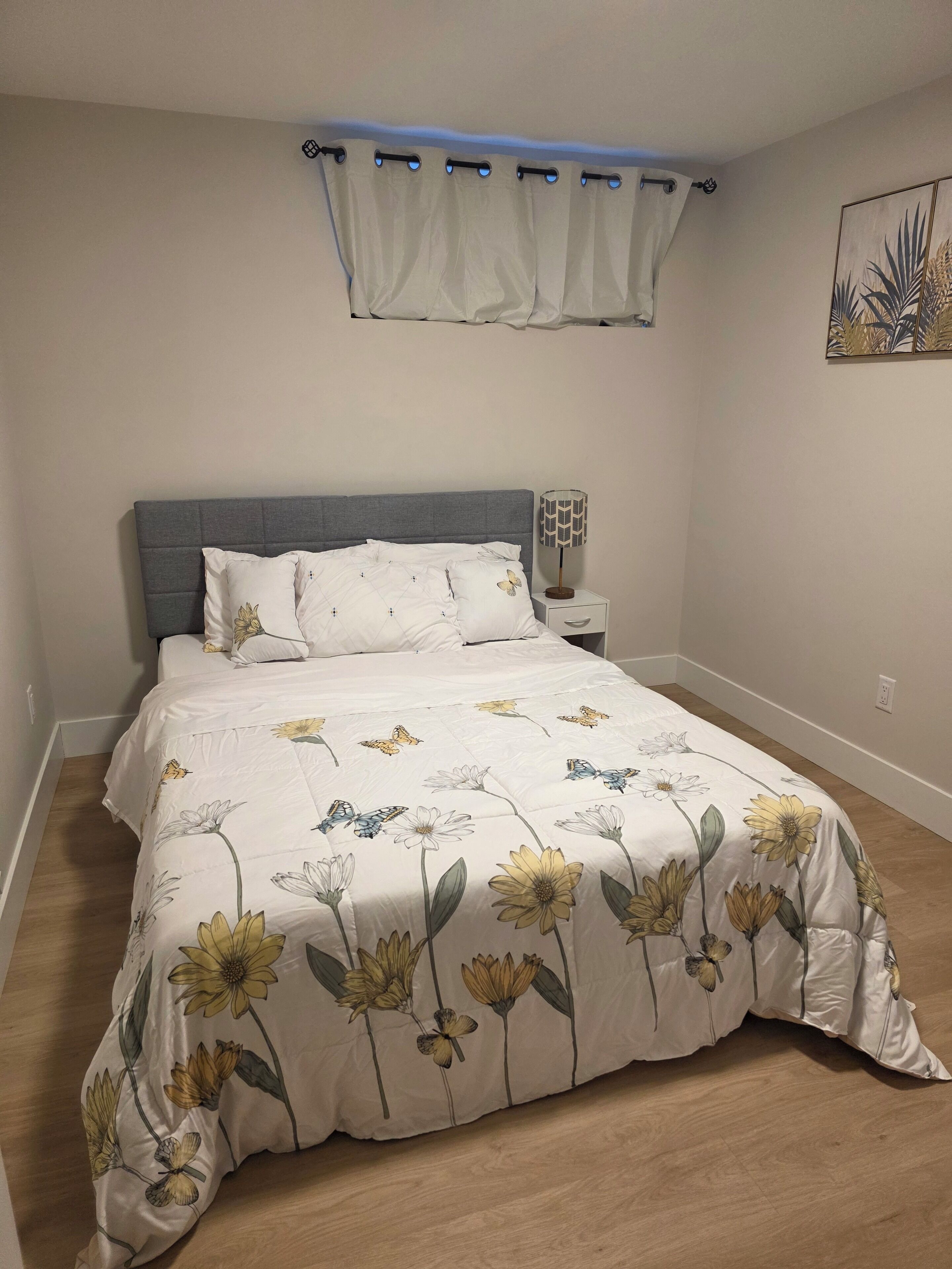 Une chambre à coucher comprenant un lit, une table de chevet, une lampe, une fenêtre avec des rideaux et une œuvre d’art accrochée au mur.