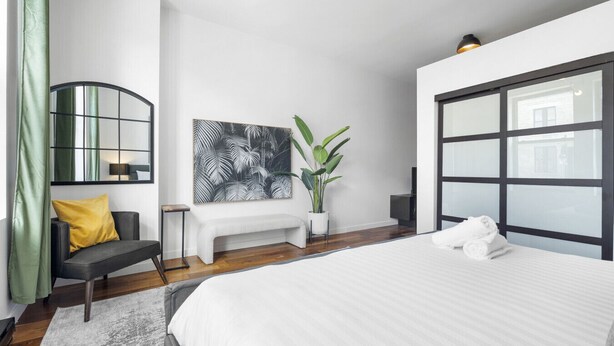 A modern bedroom with a large bed, a bench, a potted plant, and a wall-mounted mirror.