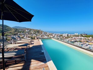 A rooftop pool with lounge chairs and an umbrella, overlooking a coastal cityscape. - Designer 2BR w/ Rooftop Pool, Gym & Stunning Views (Puerto Vallarta)