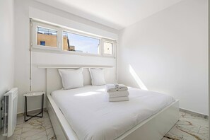Ein modernes Schlafzimmer mit einem großen Bett, einem kleinen Nachttisch, einer Heizung und einem Fenster mit Blick auf Gebäude.