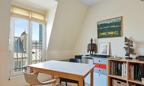 A room with a dining table, chairs, a bookshelf, a radiator, and a window with a view of buildings. - Appartement sur les toits de Paris - Saint Germain (Paris)
