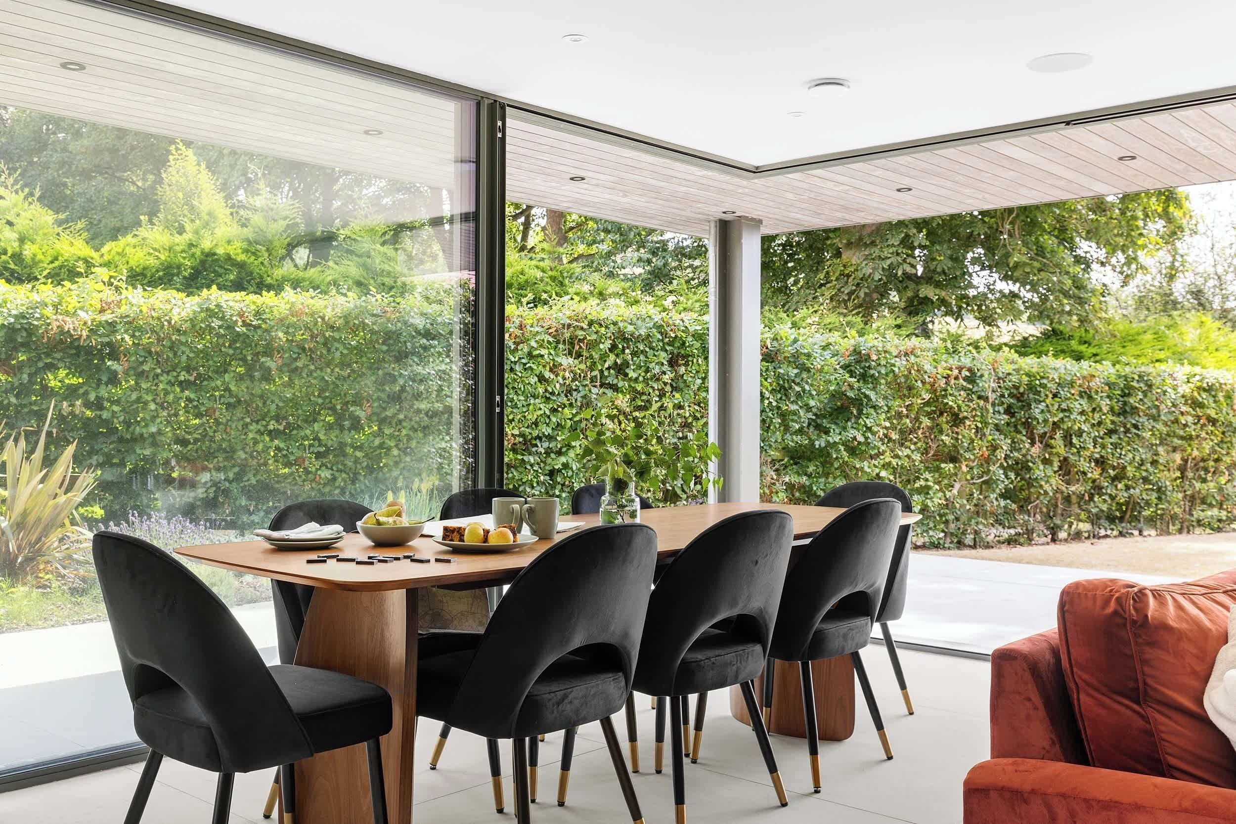 A modern dining area with a wooden table and black chairs, overlooking a lush garden.