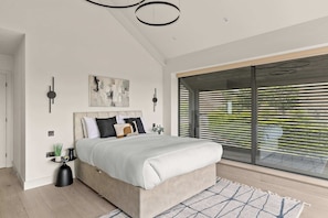 A modern bedroom with a large bed, a painting, a round wall clock, and a view through floor-to-ceiling windows.