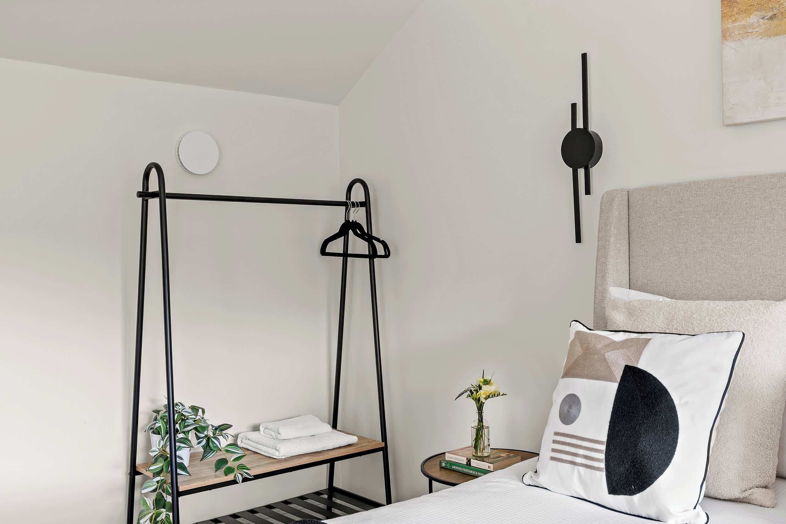 Une chambre à coucher avec un lit, une table de chevet ornée d’un vase et de livres, une lampe fixée au mur, et une étagère en métal noir sur laquelle sont posées des serviettes et des plantes.