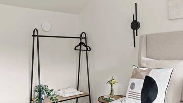 Une chambre à coucher avec un lit, une table de chevet ornée d’un vase et de livres, une lampe fixée au mur, et une étagère en métal noir sur laquelle sont posées des serviettes et des plantes.