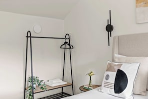 A bedroom with a bed, a bedside table with a vase and books, a wall-mounted light, and a black metal shelf with towels and plants.
