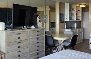 A modern hotel room with a bed, a desk with a computer, a dining table with chairs, and a kitchen area.