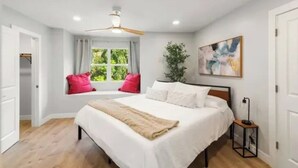 Un dormitorio con cama, ventana con cortinas, ventilador de techo, mesita de noche y una obra de arte en la pared.