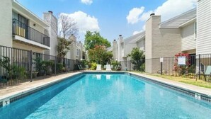 Una piscina rectangular rodeada de edificios de apartamentos con balcones, cielo despejado y una valla.