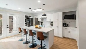 Una cocina moderna con una isla central, taburetes de bar, iluminación colgante y armarios blancos.