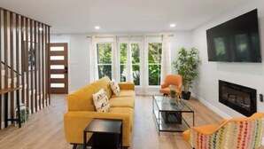 Una sala de estar con sofá, sillón, televisor, chimenea y ventanas amplias.