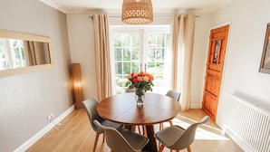 A dining area with a round table, chairs, a vase with flowers, a hanging light fixture, and a window with curtains. - Uphill Broadway (Lincoln)