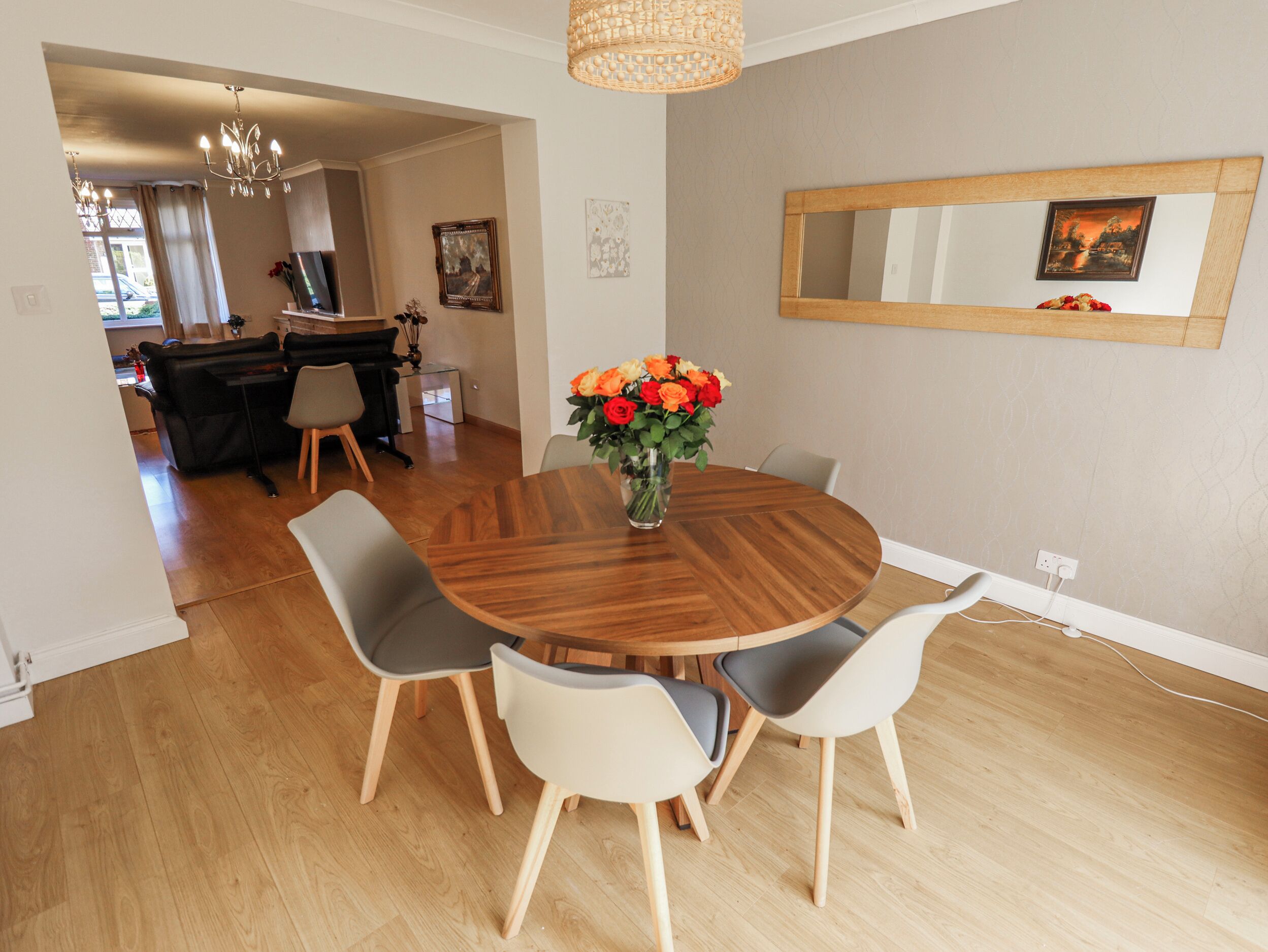 A dining area with a round wooden table, four chairs, a vase with flowers, a chandelier, and a mirror on the wall.