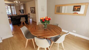A dining area with a round wooden table, four chairs, a vase with flowers, a chandelier, and a mirror on the wall.