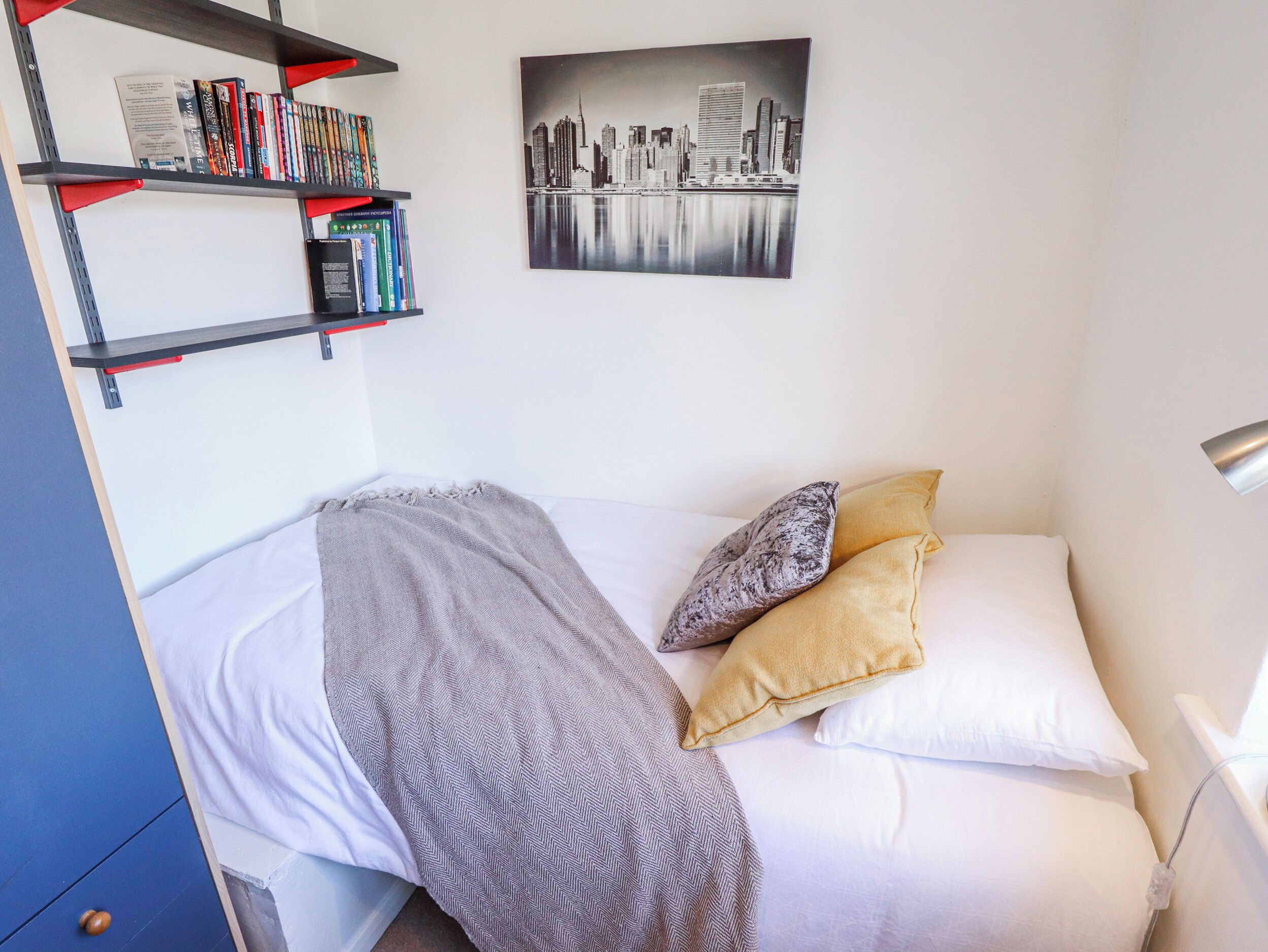 A neatly made bed with a patterned blanket and pillows, a bookshelf with books and DVDs, and a cityscape painting on the wall.
