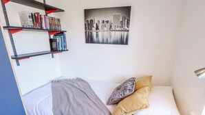 A neatly made bed with a patterned blanket and pillows, a bookshelf with books and DVDs, and a cityscape painting on the wall. - Uphill Broadway (Lincoln)