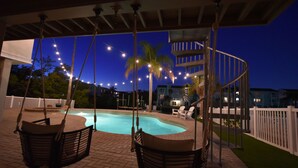 Un área de piscina con sillones colgantes, una piscina, palmeras y luces de cuerda por la noche.