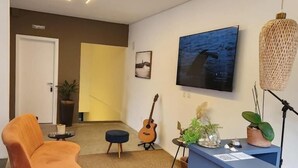 A modern living room with a sofa, a guitar, a TV, and a desk with plants. - Casa Arrô Pousada Boutique (Itajaí)