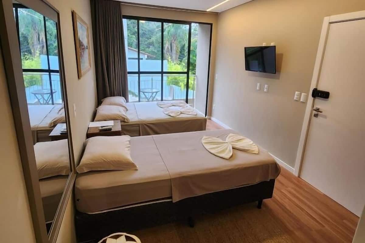 Ein Hotelzimmer mit zwei Betten, einem Fernseher, einer Tür mit Griff und einem Fenster mit Blick auf Grünflächen.