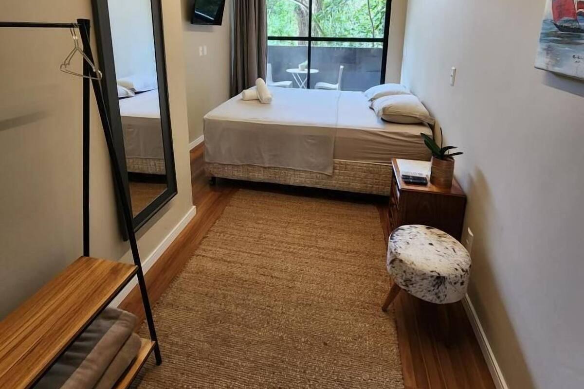 Ein Schlafzimmer mit einem Bett, einem Spiegel, einem kleinen Tisch mit Pflanze, einem Fenster mit Blick auf Grünflächen und einem Bild an der Wand.